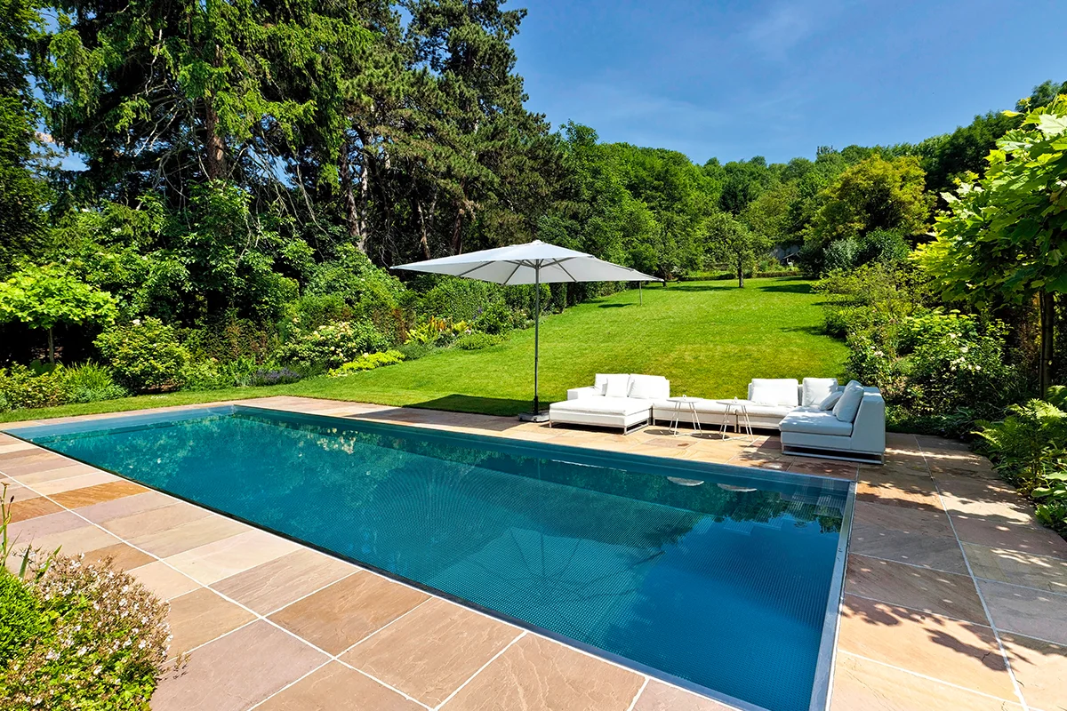 Edelstahlpool mit Natursteinterrasse, Sonnenschirm und Lounge vor einer großen Rasenfläche mit alten Bäumen