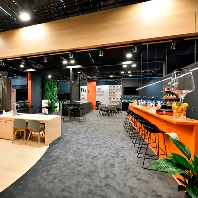 Bild in der Anzeige: Messestand mit Lounge, orangefarbener Bar und Pflanzen