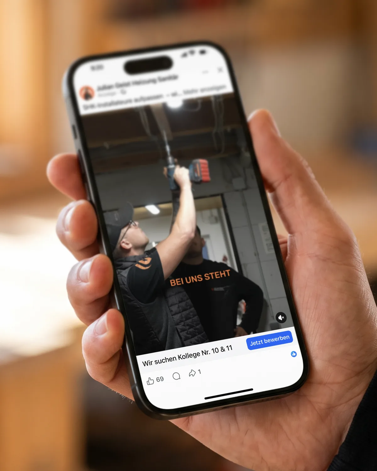 Hand hält ein iPhone mit der Facebook-Anzeige von Julian Geist Heizung Sanitär: Wir suchen Kollege Nr. 10 und 11, Knopf Jetzt bewerben